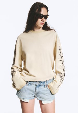Jeune personne portant des lunettes de soleil noires, un sweat-shirt beige à manches longues avec des motifs à points noirs sur les manches, et un short en jean clair.