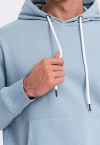 Ljusblå hoodie gjord av mjukt material, med en framficka, vita dragsnören och en avslappnad passform. Utmärkande detaljer inkluderar svarta öglor.