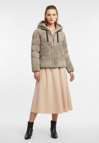 Beżowa kurtka puchowa o pionowym żebrowanym wzorze, z kapturem i czarnymi sznurkami, zestawiona z luźną brzoskwiniową spódnicą i czarnymi botkami za kostkę.