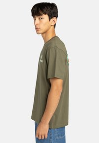 Olívovo zelené bavlnené tričko s krátkymi rukávmi. Obsahuje detail loga na prednej strane a voľný strih. Kombinuje sa s modrými džínsovými šortkami.