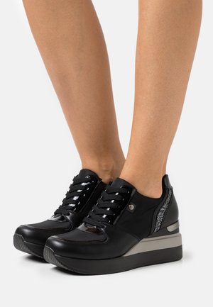 Zwarte leren sneakers met sleehak en veters aan de voorkant, glitterdetail op de hiel, gedragen op blote voeten.