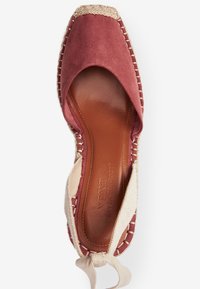 Chaussure espadrille avec une tige en suède bordeaux, une semelle intérieure en cuir brun doux et une finition en jute naturel. Présente une sangle en tissu nouée sur le côté.