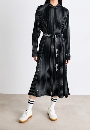 Femme portant une robe chemise midi noire à rayures blanches verticales avec une ceinture en tissu de marque, associée à des baskets blanches et des chaussettes rayées.