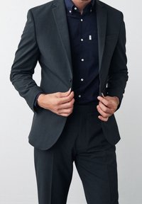 Traje gris oscuro de diseño ajustado, blazer de un solo botón y pantalones a juego. Debajo, una camisa azul marino con botones blancos.