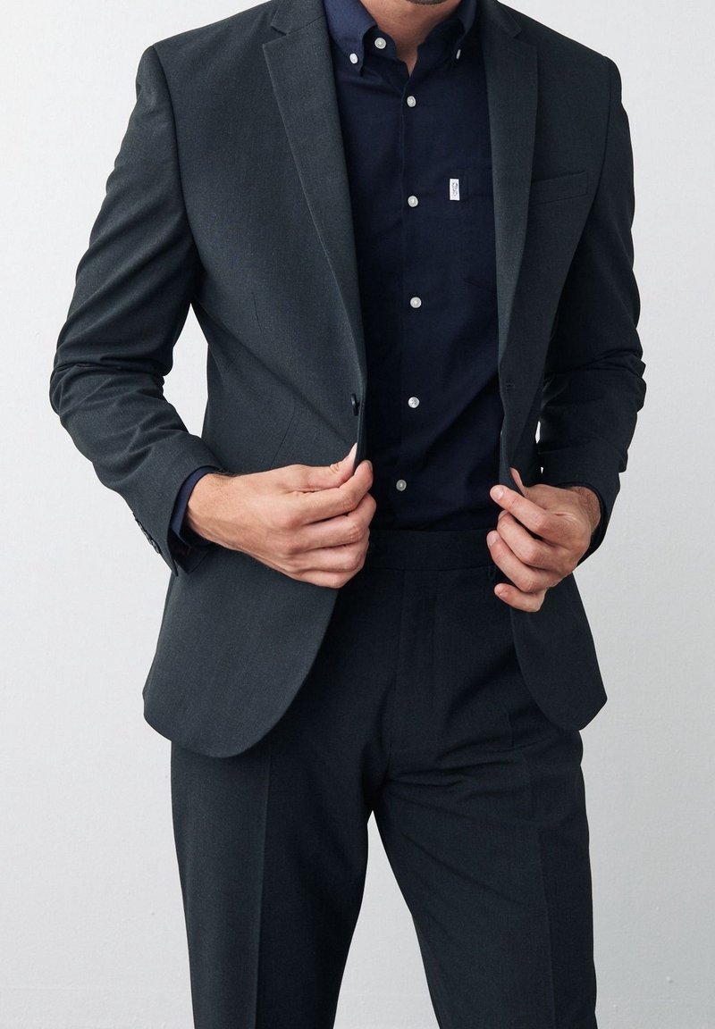Traje gris oscuro de diseño ajustado, blazer de un solo botón y pantalones a juego. Debajo, una camisa azul marino con botones blancos.