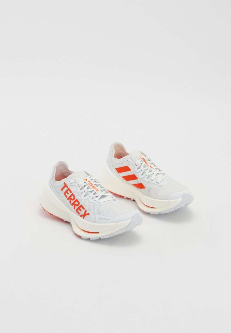 White running shoes with orange accents. Mesh material, rounded toe, and thick midsole. "TERREX" printed on lateral side and three stripes on heel.