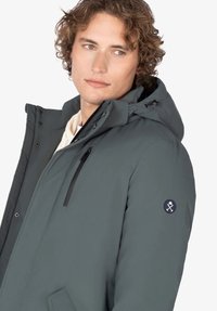 Joven con cabello rizado que lleva una chaqueta de invierno con capucha gris, con un bolsillo con cierre en el pecho y un parche de logo en la manga.