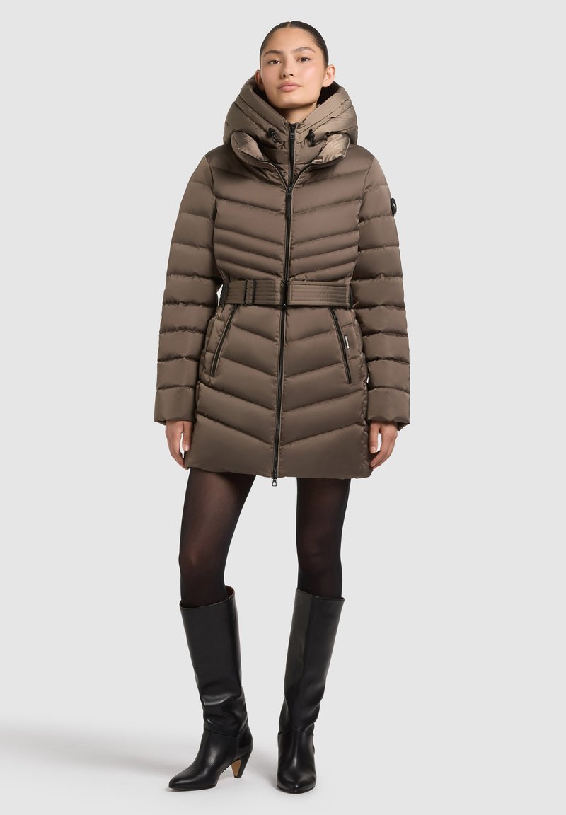 Hnedá nadýchaná bunda s prešitým vertikálnym vzorom, sťahovaným pásom a kapucňou; obsahuje zipsy a je skombinovaná s koženými čižmami po kolená.