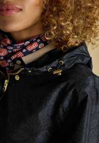 Giacca nera con motivo floreale, accessori dorati e cappuccio con coulisse. Sciarpa rosa e blu con motivo visibile sotto.