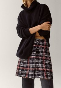 Zwarte oversized trui met een hoge kraag, gecombineerd met een geruite rok in zwart, wit en rood. Soepele texturen en een losse pasvorm.