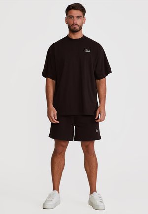 Mann står, iført svart oversized T-skjorte og matchende shorts med hvite joggesko, ensfarget lys bakgrunn.