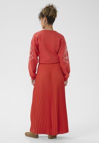 Sieviete ar šķipsnām matiem, ģērbusies zelta riņķa auskaros, sarkanā džemperī ar baltu ziedu izšuvumu uz piedurknēm, garā sarkanā plandošā svārkā un brūnās kurpēs.