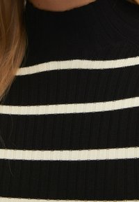 Haut noir à col montant côtelé avec des rayures blanches horizontales. La texture est tricotée, créant une silhouette ajustée.