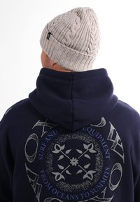 Bonnet en tricot beige avec un motif de câble texturé. Sweat à capuche bleu marine foncé avec une grande impression graphique dans le dos, comportant des motifs circulaires et floraux.