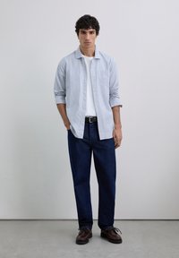 Camisa de botón a rayas azul y blanca con mangas largas, combinada con jeans azul oscuro y zapatos marrones de cordones. Ajuste simple y relajado.