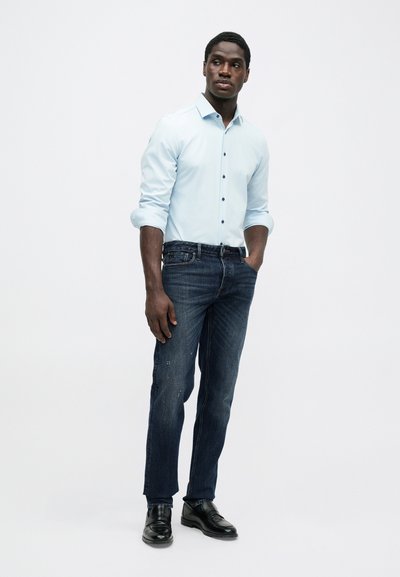 Mann står med en hånd i lommen og har på seg lys blå skjorte med oppbrettede ermer, mørk blå jeans og svarte pensko på hvit bakgrunn.