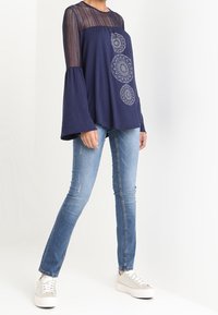 Blusa azul marino con espalda de encaje transparente, mangas acampanadas y detalles de bordado circular, combinada con pantalones vaqueros azules y zapatillas blancas.