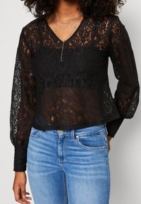 Černá krajková blůza s dlouhými nařasenými rukávy a výstřihem do V, s květinovým vzorem a průhlednou texturou, zkombinovaná se světle modrými džíny.