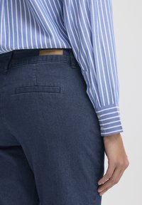 Világoskék-fehér csíkos hosszú ujjú ing sötétkék farmernadrággal, mely hátsó zsebbel és világosbarna bőr díszítéssel a derékrészen.