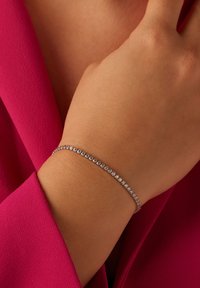 Zilveren armband met ronde heldere stenen, gedragen om een pols. De achtergrond heeft een helder roze stof. De gladde textuur versterkt de glans.