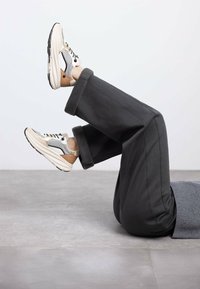 Sneaker con una toma in rete beige e bianca, lacci neri e suola ammortizzata; pantaloni grigi a gamba larga con risvolti appoggiati su un pavimento grigio.