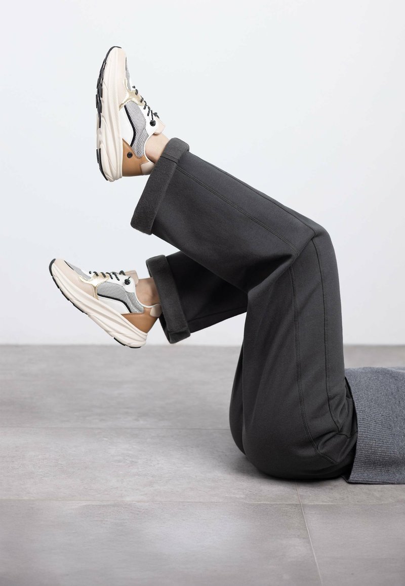 Sneaker con una toma in rete beige e bianca, lacci neri e suola ammortizzata; pantaloni grigi a gamba larga con risvolti appoggiati su un pavimento grigio.