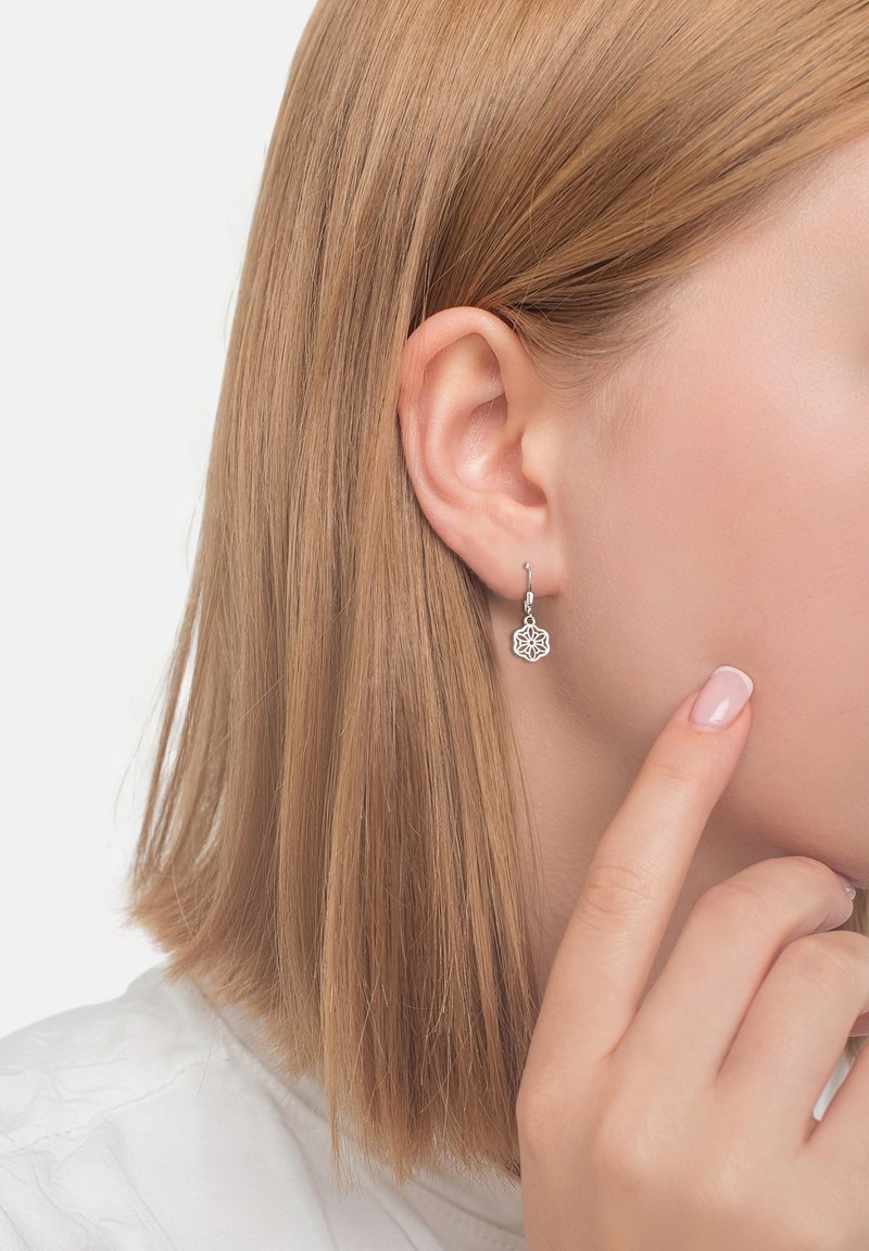 Gros plan d'une personne aux cheveux blonds raides portant une petite boucle d'oreille pendante florale en argent à l'oreille gauche, avec un doigt touchant le visage.