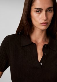 Femme aux longs cheveux foncés portant un polo noir texturé à manches courtes avec boutons, regardant devant elle avec une expression neutre.