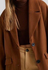 Brązowa, wełniana, oversize'owa marynarka z pod spodem czekoladowym golfem o prążkowanej fakturze. Czarne guziki i minimalne przeszycia na kieszeniach.