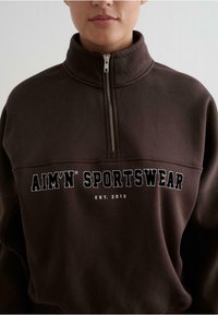 Sudadera de forro polar marrón con un alto cuello y cierre de media cremallera. Presenta un logotipo bordado en blanco que dice "AIM’N SPORTSWEAR EST. 2013" en el frente.