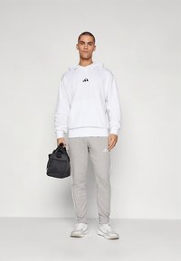 Λευκή φούτερ με κουκούλα και μαύρο λογότυπο, γκρι φόρμα με λευκές ρίγες και μαύρη τσάντα. Λεπτή εφαρμογή, λείο ύφασμα, casual αθλητικό στυλ.