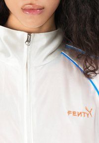 Giacca leggera bianca con zip, dettagli blu e logo ricamato arancione "FENTY" sul petto, dotata di colletto alto e tessuto texturizzato.