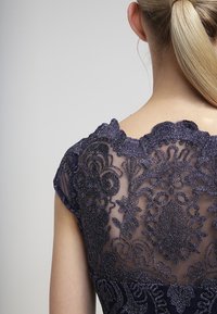 Tummansininen mekko, jossa on läpikuultava pitsiyksityiskohta takana, sisältäen monimutkaisia kuvioita ja tekstuurisia koristeita; lyhyet hihat ja kaareva kaula-aukko.