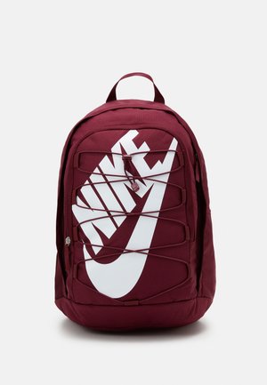 Sac à dos marron avec grand logo Nike blanc stylisé, poche avant zippée et cordons élastiques réglables sur le panneau avant.