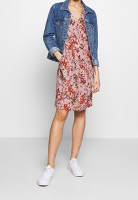 Kukkamekko vaaleanpunaisena, punaisilla ja valkoisilla kuvioilla, täydentää farkkutakki. Mallilla on valkoiset tennarit rennon ilmeen luomiseksi.