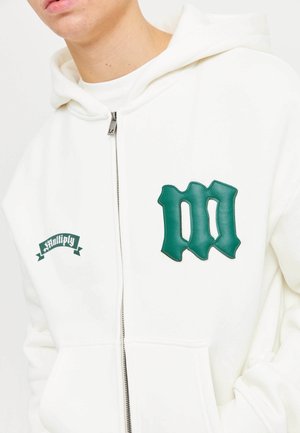 Person iført en hvid zip-up hoodie med et grønt stiliseret "m" patch og små "multiply" tekster på brystet, hænderne i lommerne.