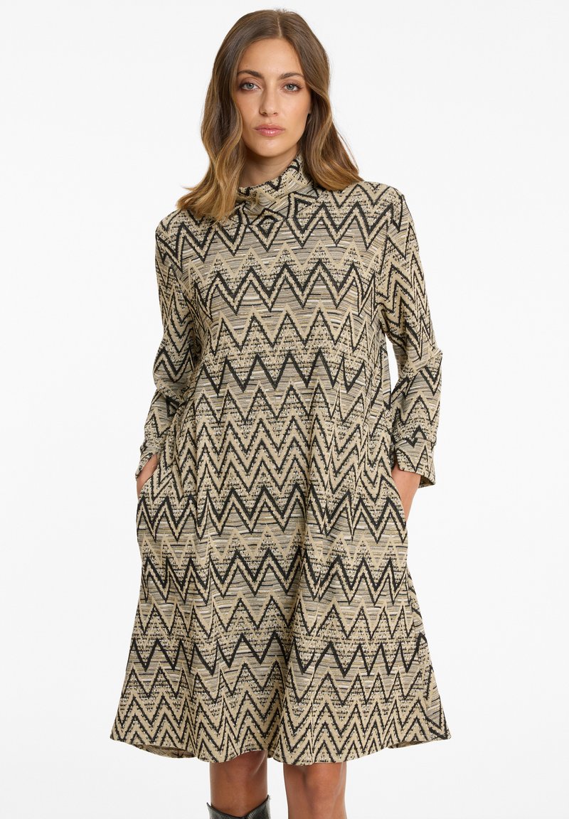 Frau in einem knielangen beigen Kleid mit schwarzen Zickzackmustern, langen Ärmeln, hohem Kragen und Händen in den Taschen, vor einem weißen Hintergrund.