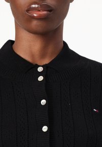 Svart strikket cardigan med ribbet krage, med fire perleknapper og perforert tekstur. Liten merke-logo i rødt, hvitt og blått.