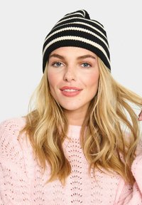 Černo-beige pruhovaná pletená čepice s špičatým vrcholem. Modelka má na sobě růžový svetr z otevřené pleteniny s jemným vzorem.