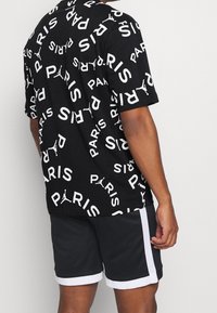 Mężczyzna w czarnej koszulce z białym napisem "PARIS" oraz logo Jumpman, połączonej z czarnymi szortami sportowymi z białymi paskami po bokach i na dole.