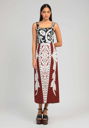 Jeune femme debout, portant une longue robe sans manches à motifs floraux et géométriques noirs, blancs et rouges, avec des sandales à plateforme noires.