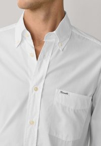 Uomo che indossa una camicia bianca con bottoni, colletto abbottonato e una tasca sul petto con la scritta "Façonnable".