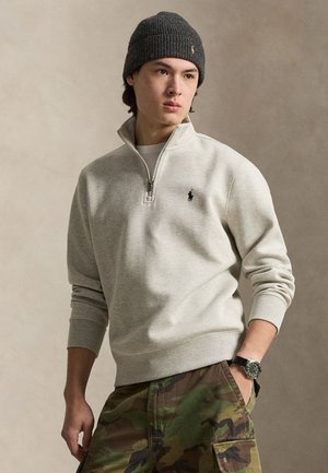 Młody mężczyzna w szarej czapce typu beanie, jasnoszarej bluzie z suwakiem do ćwiartki, spodniach w kamuflaż i zegarku na rękę, stojący z jedną ręką w kieszeni.