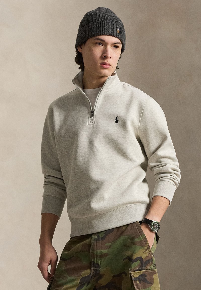 Jonge man met een grijze beanie, lichtgrijze trui met een kwart rits, camouflagebroek en een polshorloge, staand met één hand in de zak.
