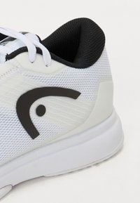 Hvide sportssko med en åndbar mesh-overdel, sorte accenter og et struktureret design. Har et rundt logo nær snørebåndene.