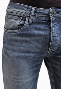 Tummansiniset farkut haalistuneella pesulla, viisipokkoinen malli, metalliset yksityiskohdat ja näkyvät tikkauskoristukset vyötäröllä ja taskuissa.