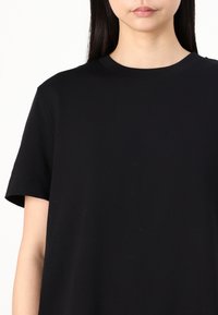 Femme portant un t-shirt noir uni à manches courtes et col rond, visible des épaules au milieu de la poitrine sur fond blanc.