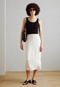 Top sin mangas negro con botones, falda midi blanca y fluida, sandalias negras deslizadas y un pequeño bolso negro sobre un fondo neutro.