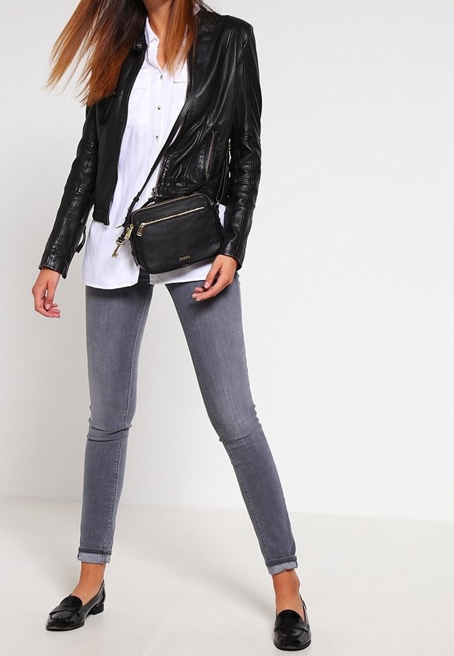 Frau, die eine schwarze Lederjacke, ein weißes Shirt, graue Skinny-Jeans, schwarze Loafer trägt und eine schwarze Umhängetasche hält, steht vor einem weißen Hintergrund.