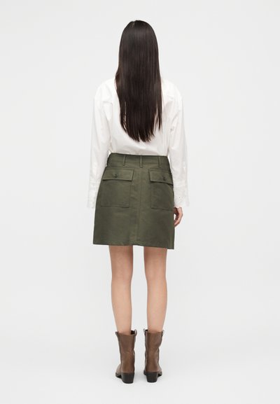 Φούστα σε ελιά πράσινο με δύο πίσω τσέπες, υφή υφάσματος και εφαρμοστή μέση. Συνδυασμένη με λευκό πουκάμισο και καφέ μποτάκια μέχρι τον αστράγαλο.